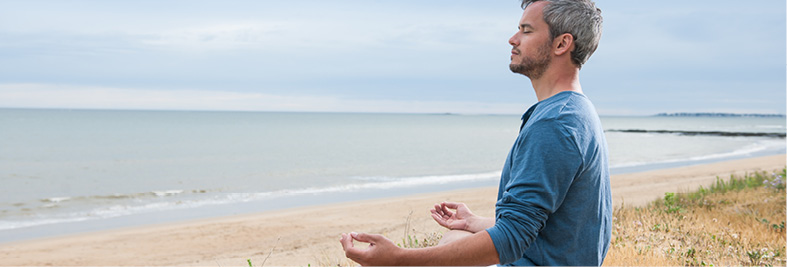 Man with short grey hair sits with eyes closed and hands resting on his knees in a meditative pose beside the calm ocean.