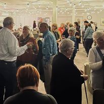 A diverse crowd of people stands together in a large, well-lit hall, mingling and talking in a busy indoor social space.