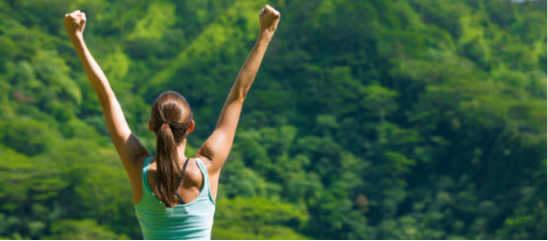 A woman stands with her back to the viewer, arms raised high toward the lush green mountain foliage in a joyful stance.