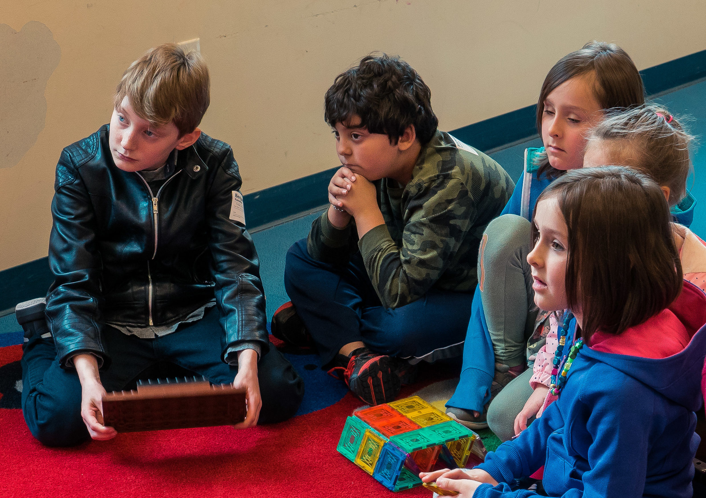 Five children sit together on a bright red carpet, focused on a colorful building toy and a block held during a lesson.