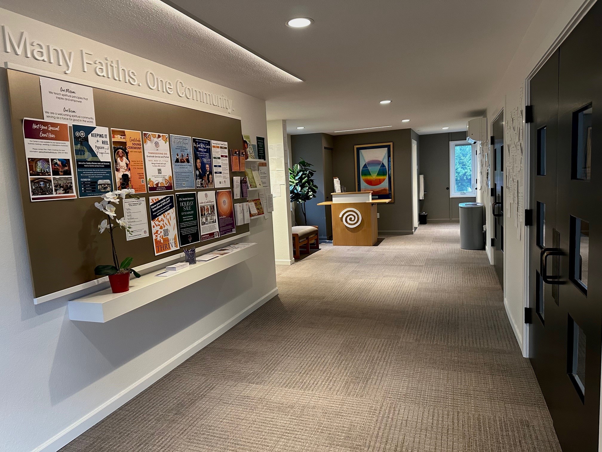 Community bulletin board with rows of flyers lines the wall of a bright carpeted hallway leading to a wooden welcome desk.