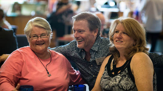A man in a patterned shirt smiles while resting his arm on the shoulders of two women seated beside him at a cafe table.