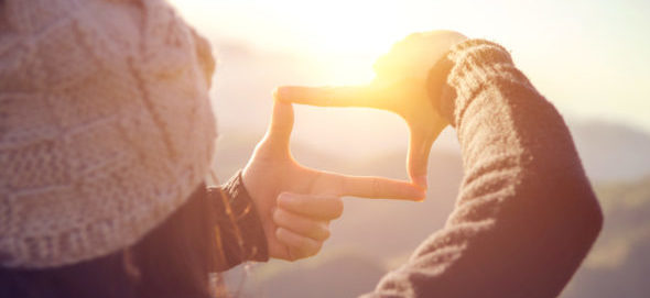 Bright sunlight glows intensely behind two fingers framing the distance, casting a warm, hazy lens flare across the view.