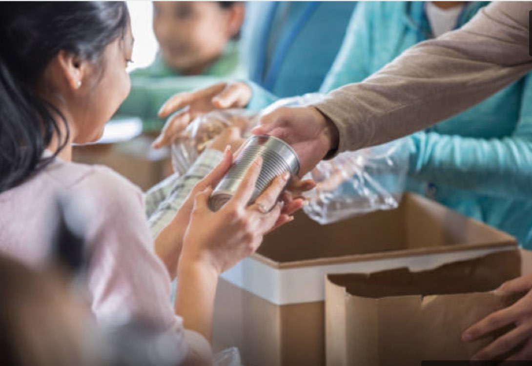 Young girl carefully places a metal food can into a cardboard box while a volunteer helps her sort donations at a table.